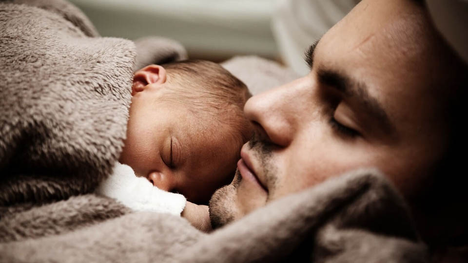 A man cradles a warmly wrapped baby as they both sleep